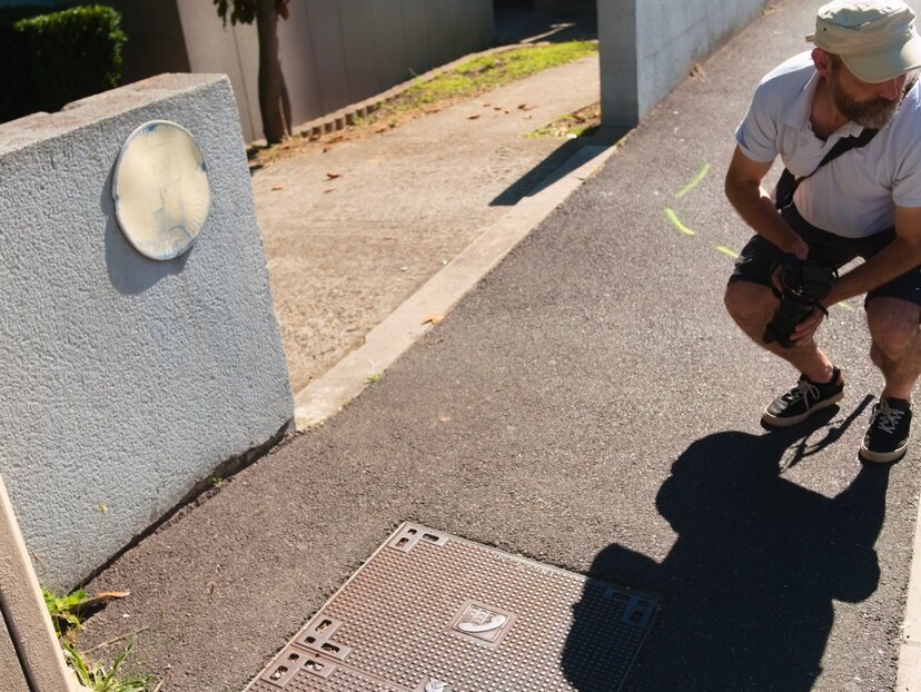 Photo du photographe accroupi, dans la recherche du bon angle, au-dessus d'une plaque en fonte télécoms, sur le trottoir, en plein soleil 